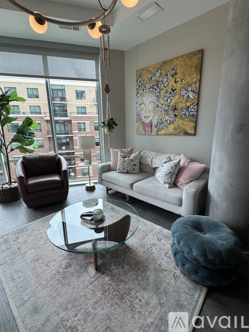 A living room with a grey couch, a glass coffee table, a brown leather chair, a blue pouf, a large painting on the wall, and a window with a view of a building outside.
