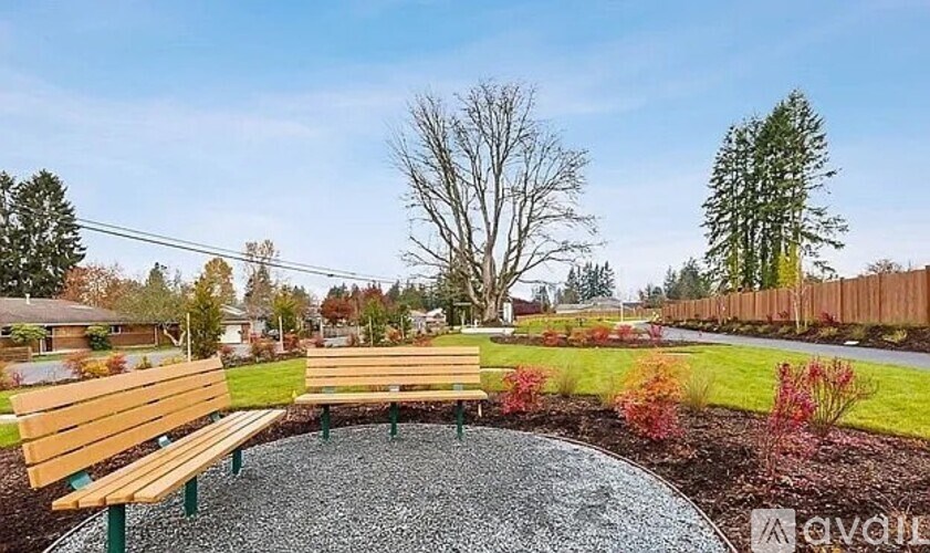 A park bench sits in the middle of a gravel area with a grassy area and trees in the background.