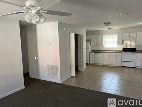 A spacious living room with a ceiling fan and a kitchen area in the background.