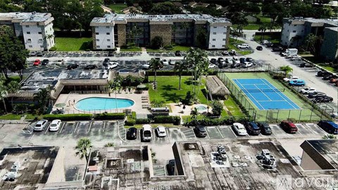 A tennis court is surrounded by a parking lot and a pool.