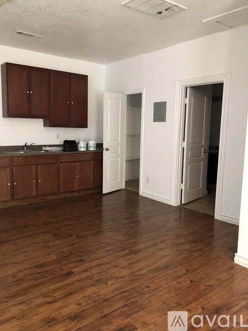 A room with wooden floors and white walls, with a kitchen area to the left and an open doorway to another room to the right.