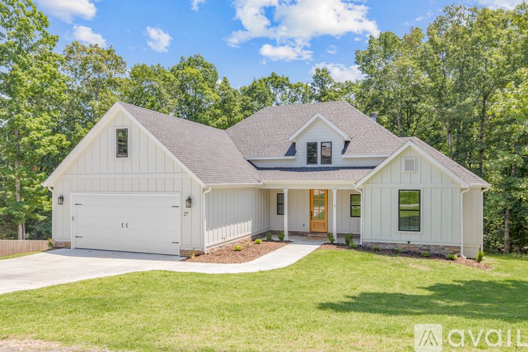 A white house with a brown roof and a brown door is surrounded by greenery.