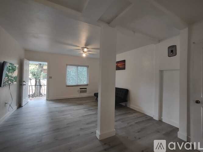 A spacious room with wooden floors and white walls, featuring a ceiling fan and a window with a view of trees.