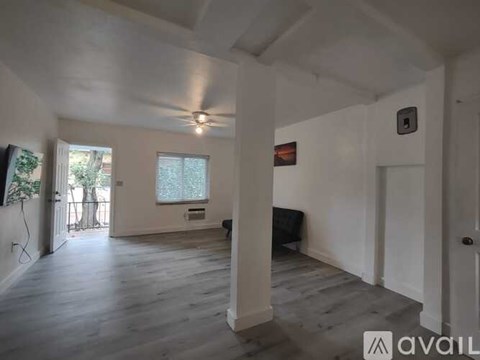 A spacious room with wooden floors and white walls, featuring a ceiling fan and a window with a view of trees.