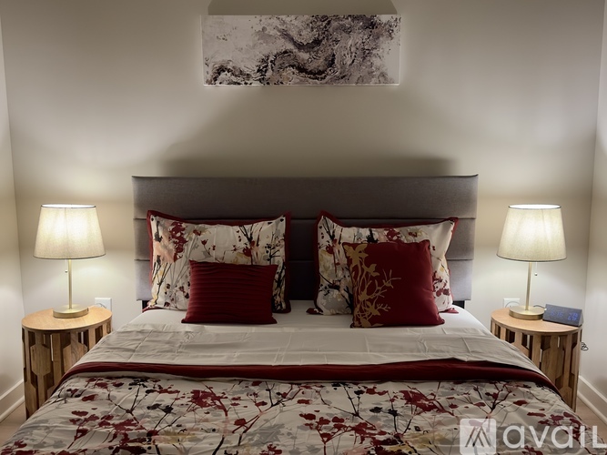 A bed with a floral patterned comforter and two red pillows is flanked by two small tables with lamps on them.