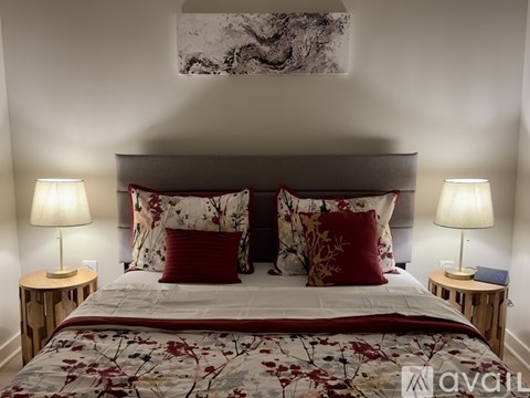 A bed with a floral patterned comforter and two red pillows is flanked by two small tables with lamps on them.