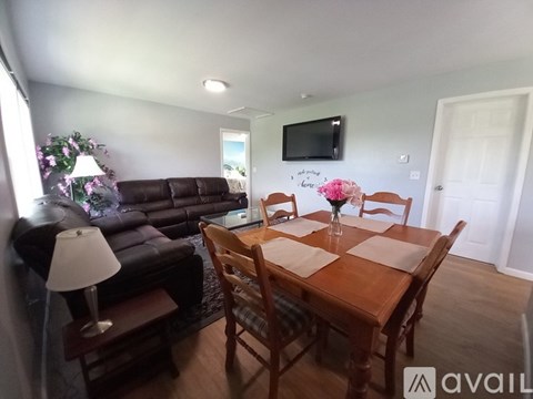 A living room with a brown leather couch, a wooden dining table with chairs, and a flat-screen TV mounted on the wall.