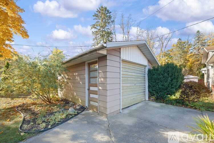 A small detached house with a garage door is surrounded by trees and shrubs.