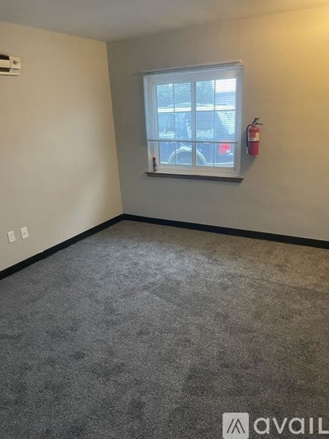 A room with a grey carpet and a window with a fire extinguisher on the wall.