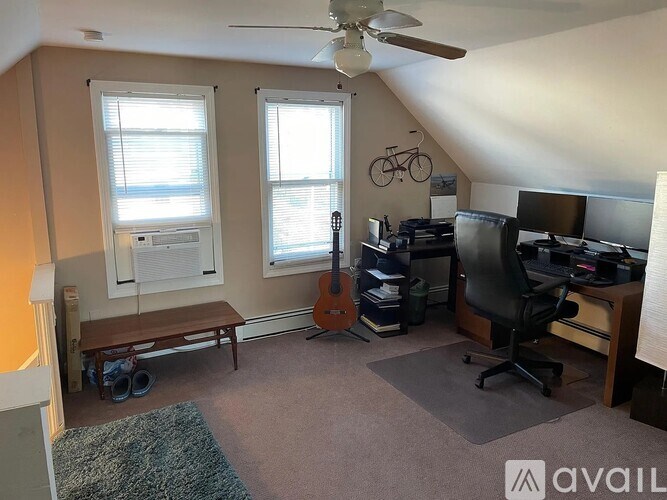 A room with a ceiling fan, a desk with a computer, a chair, a guitar, and a window with blinds.