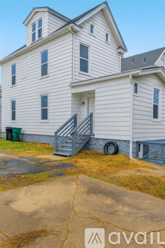 A white house with a grey roof and a green trash can in front of it.