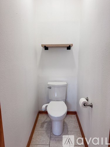A white toilet in a small bathroom with a wooden shelf above it.