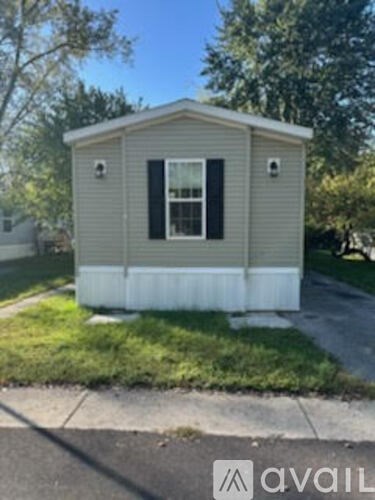 A small house with a grey roof and black shutters is situated on a grassy area.