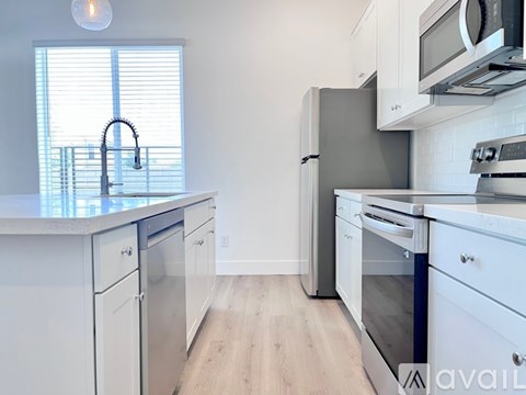 A kitchen with white cabinets and appliances, a window with blinds, and a wooden floor.