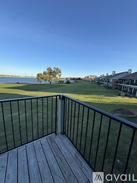 A balcony with a wooden floor and a metal railing overlooks a grassy area with a tree and a building in the distance.