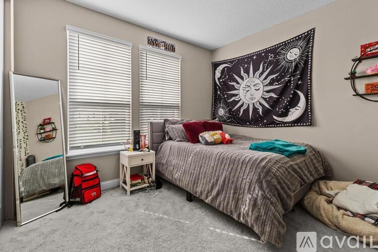 A bedroom with a bed, a desk with a chair, a window with blinds, a wall hanging, and a mirror.