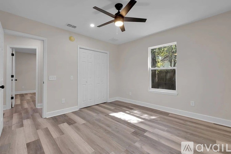 A room with a ceiling fan and light fixture, wooden flooring, and a window with a view of trees.