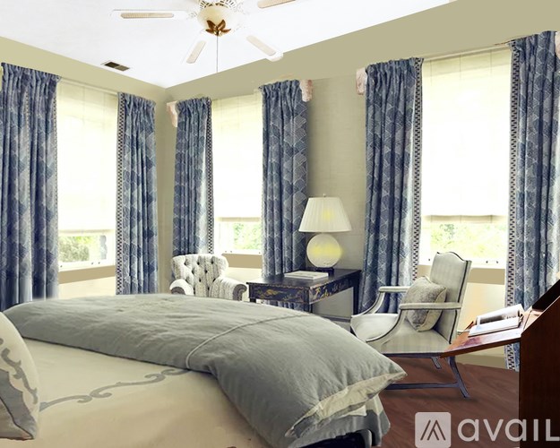 A bedroom with a large bed, a chair, a desk with a lamp, and a ceiling fan.