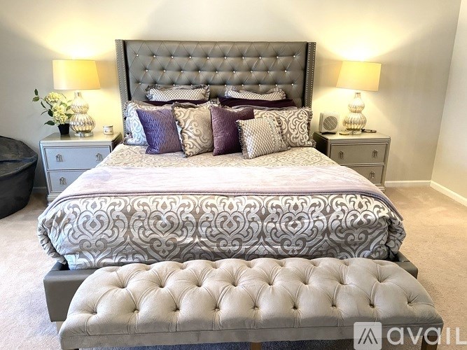 A bedroom with a large bed and a tufted ottoman in the foreground.
