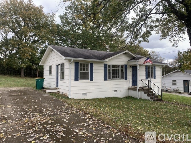 A white house with a black roof and a green trash can in front.