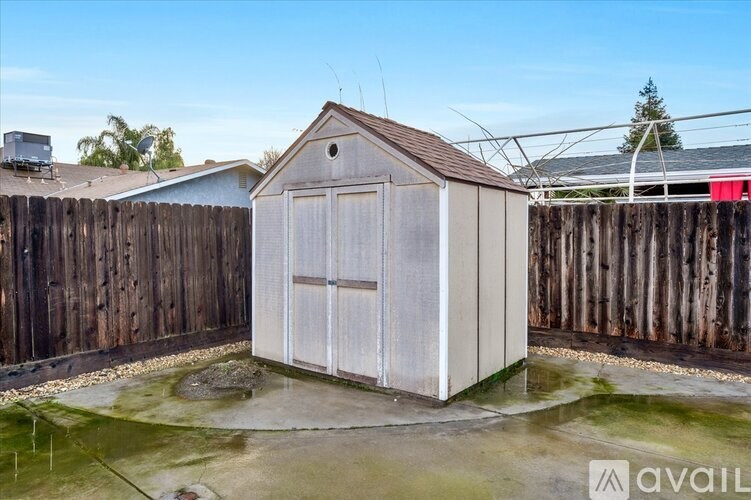 A small, white outhouse with a brown roof is situated in a backyard with a wooden fence.