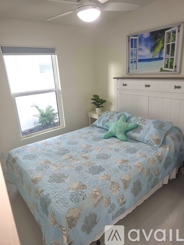 A bed with a blue and white patterned comforter sits in a room with a window and a painting on the wall.