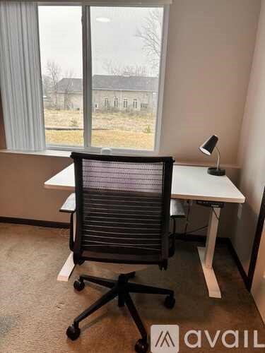 A black office chair is in front of a desk with a lamp and a window showing a view of a field and a building.