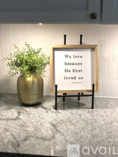 A framed sign that says "We love because He first loved us" is on a stand next to a plant.