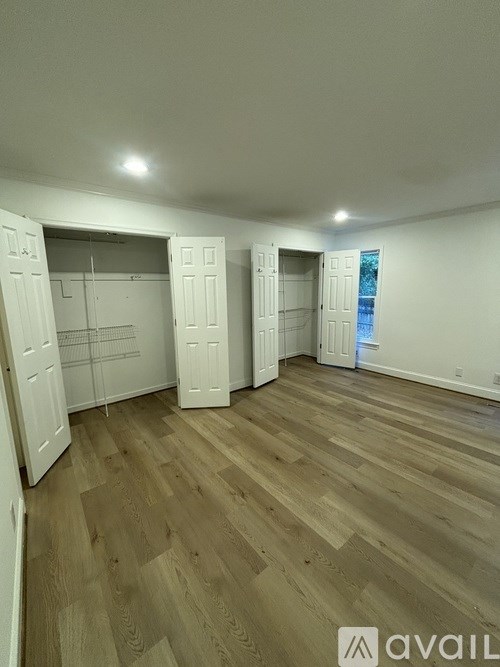 A room with wooden flooring and white walls, with a door on the left and a window on the right.