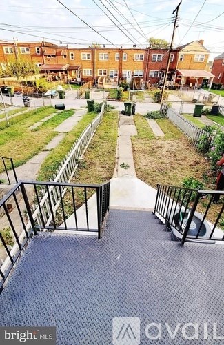 A balcony with a metal railing and a textured floor leads to a grassy area with a pathway and a fence.