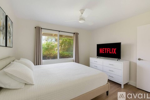 A bedroom with a bed, a dresser with a Netflix sign on it, and a window with a view of trees.