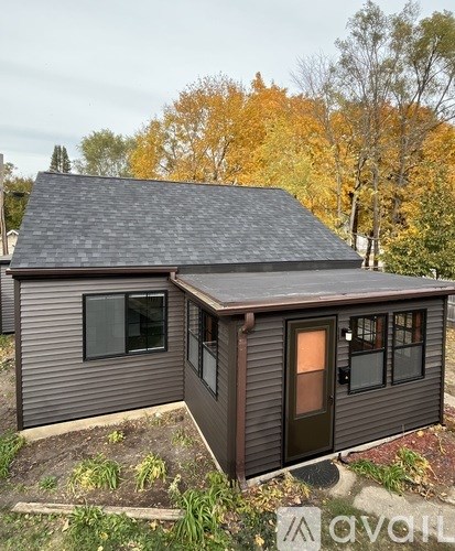 A small house with a brown door and windows is surrounded by trees with yellow leaves.