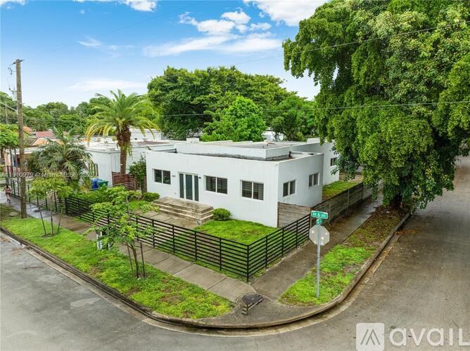 A white house with a green fence is surrounded by greenery.