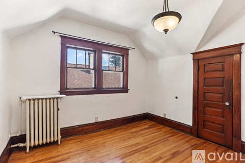 A room with a wooden floor, a window with a view of a roof, a ceiling light, and a wooden door.