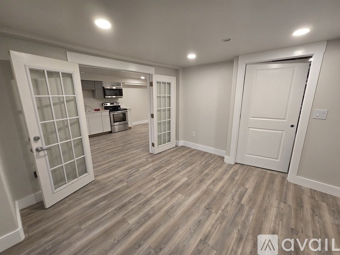 A room with white walls and wooden flooring, with a kitchen visible through a glass door.