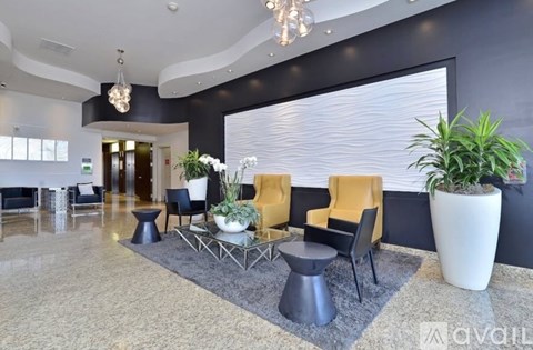 A spacious living room with a grey floor, a large window with blinds, and a variety of furniture including a yellow armchair, a black chair, and a white vase with a plant.