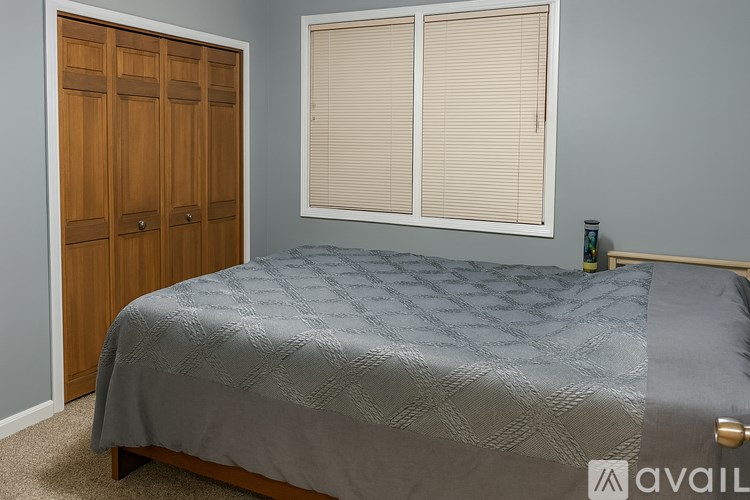 A bed with a grey comforter is in a room with a wooden wardrobe and a window with blinds.