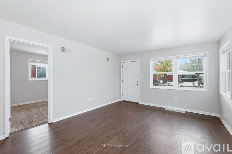 A room with wooden floors and white walls, with a window showing a view of trees and houses.