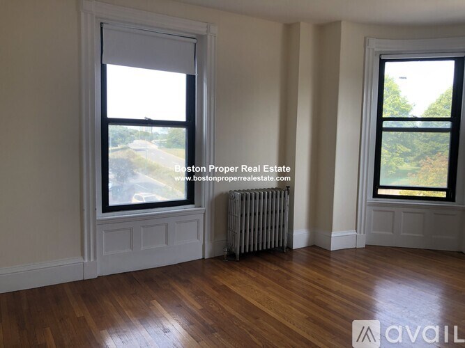 A room with wooden floors and two windows, one of which has a view of a tree-lined street.