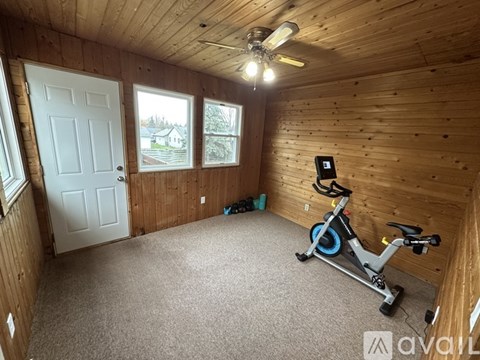 A room with a wooden ceiling and walls, a white door, a window with a view of a house, a carpeted floor, a stationary exercise bike, and a ceiling fan.