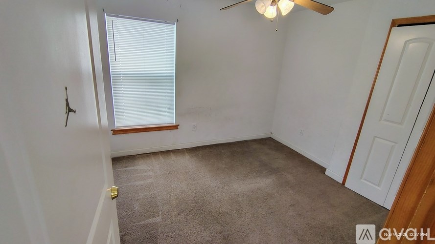 A room with a carpeted floor, a ceiling fan, and a door leading to another room.