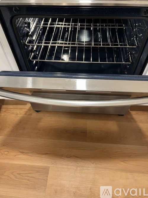 A modern oven with a stainless steel finish and a glass door is placed on a wooden floor.