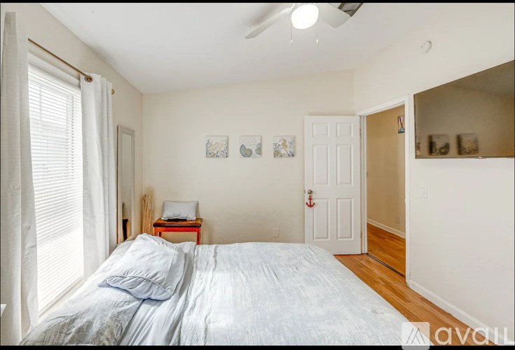 A bedroom with a bed, a window with curtains, a ceiling fan, and a small table with a lamp.