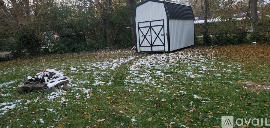 A white shed sits in a grassy field with a pile of wood in front of it.