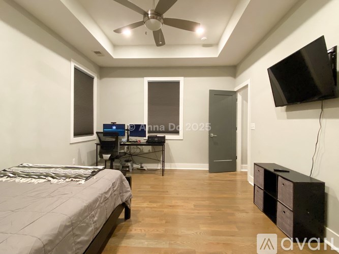 A bedroom with a bed, a desk, a ceiling fan, and a television mounted on the wall.