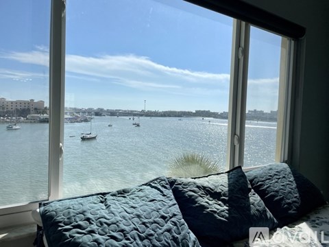 A bedroom with a large window overlooking a body of water with boats and a city skyline.