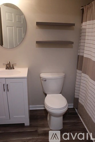 A white toilet in a bathroom with a round mirror and shelves above it.