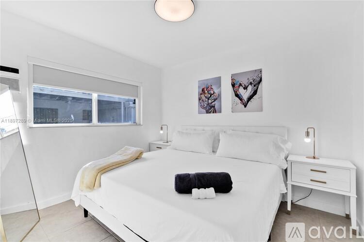 A bedroom with a white bed and a small table with a lamp on it.