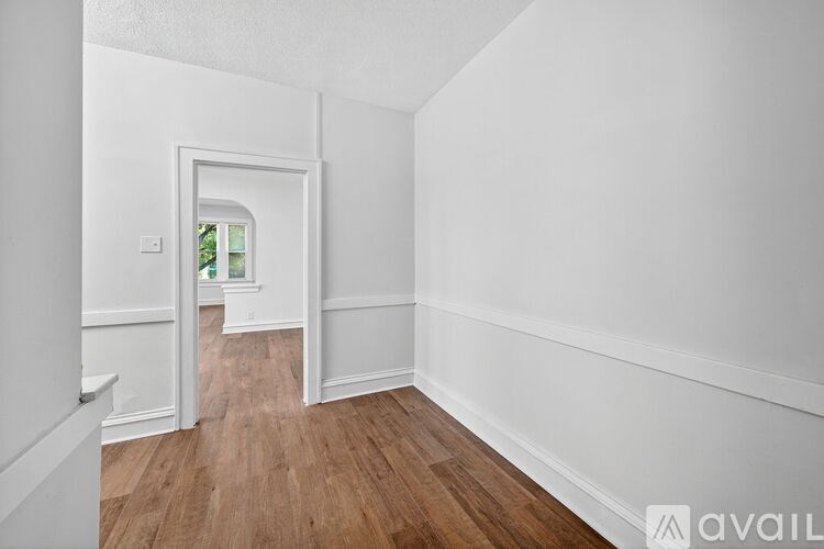 A room with white walls and wooden flooring, leading to a hallway.