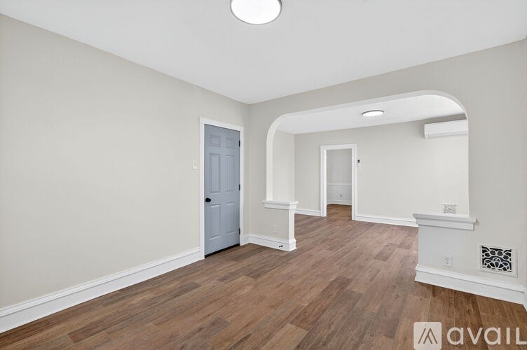 A room with a light wooden floor and white walls, with a door on the left and a window on the right.